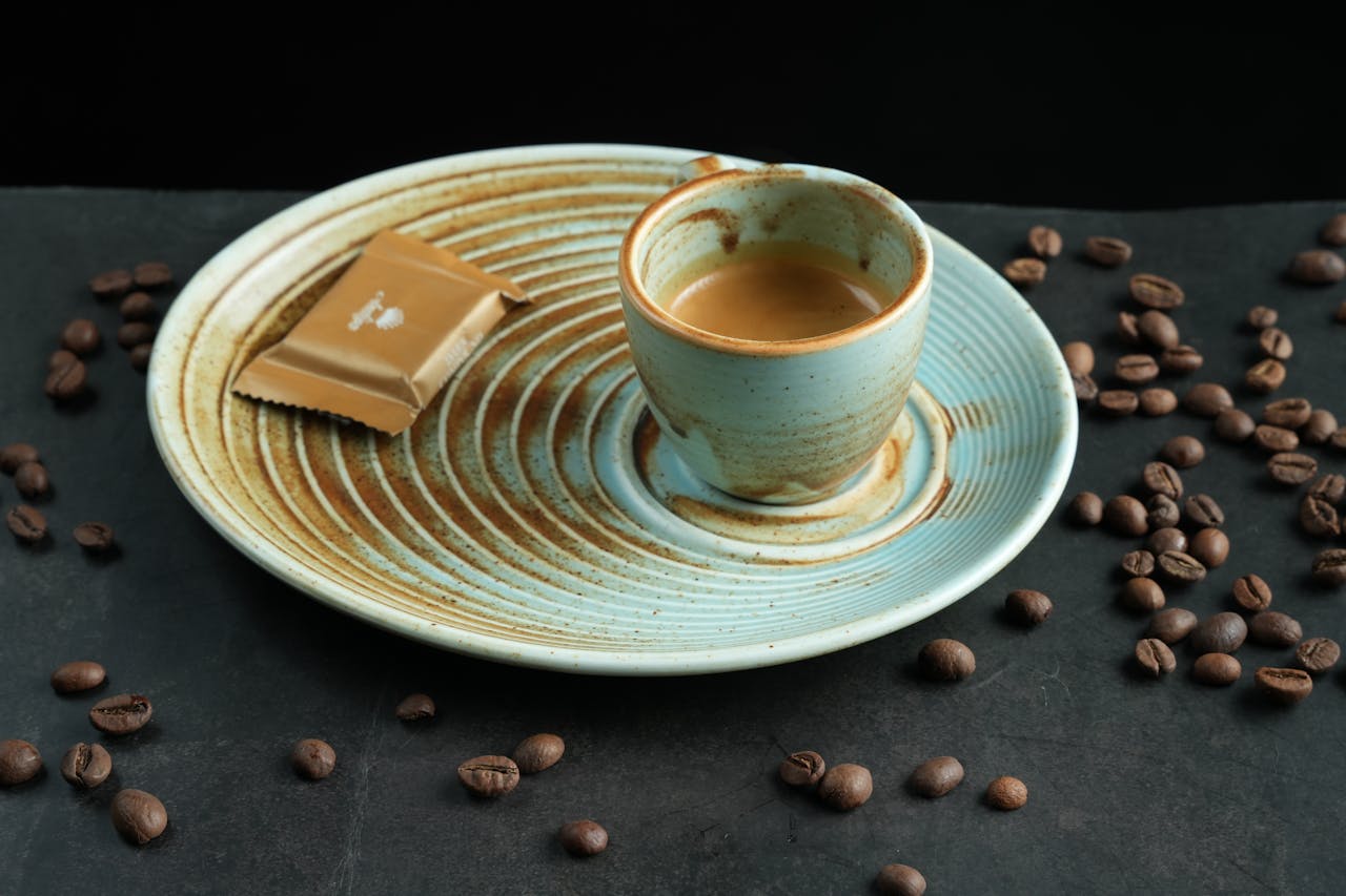 A stylish espresso coffee shot with scattered coffee beans and a chocolate biscuit on a ceramic plate.