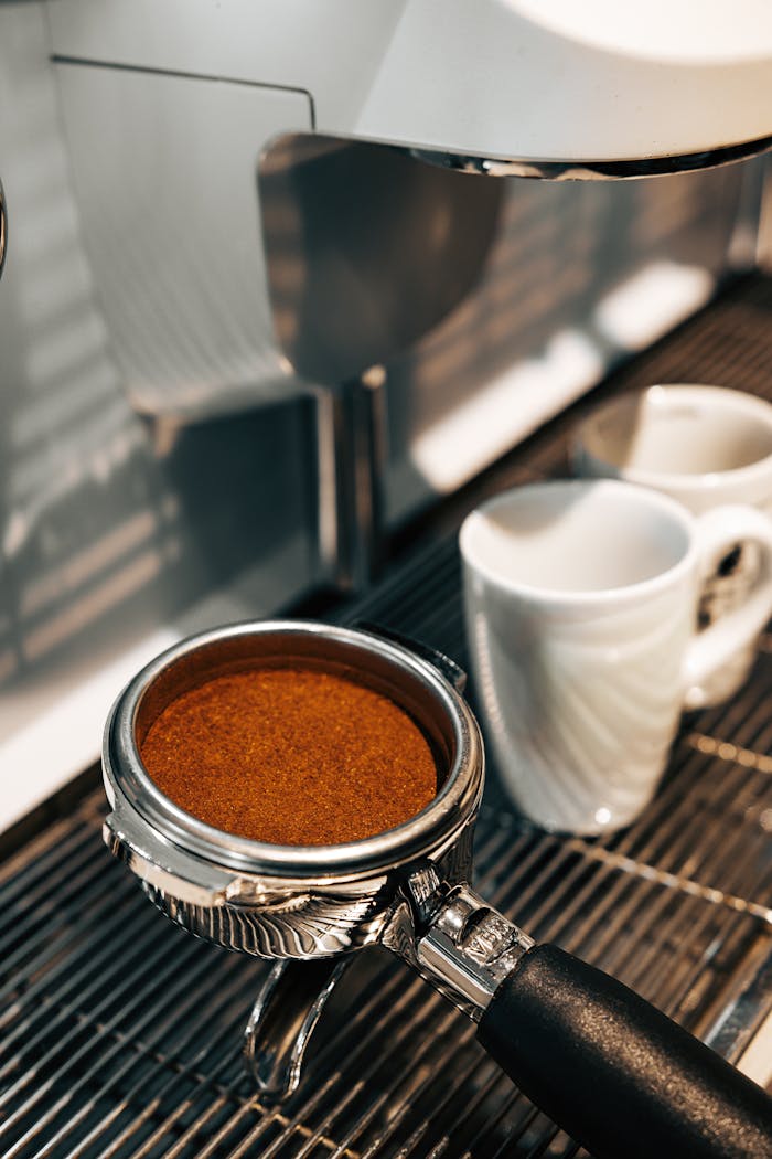 Close-up of a portafilter filled with ground coffee next to mugs at a café.