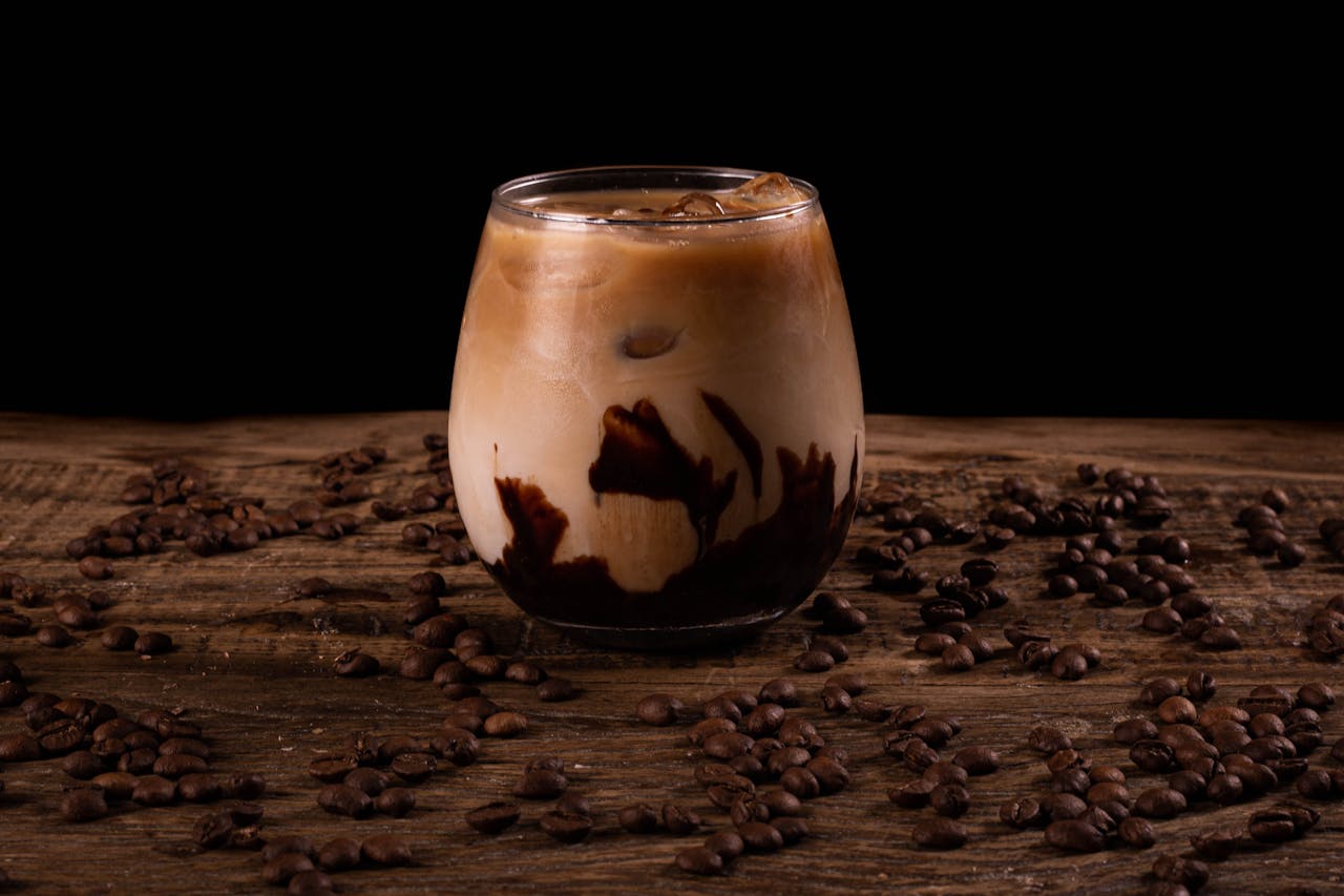 A glass of creamy iced coffee with swirls on a rustic wood table surrounded by coffee beans.